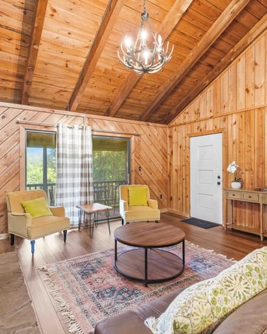Wooden cabin interior with vaulted ceiling, antler chandelier, yellow armchairs, round coffee table, and glass doors opening to deck
