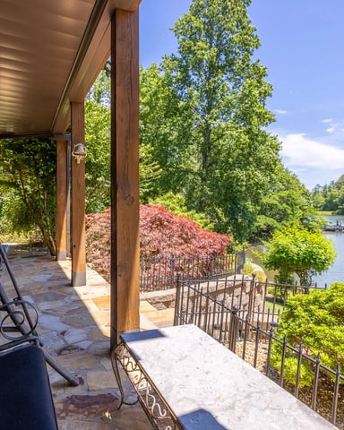 Covered porch with blue swing chairs overlooking lake, flowering shrubs, and forested shoreline