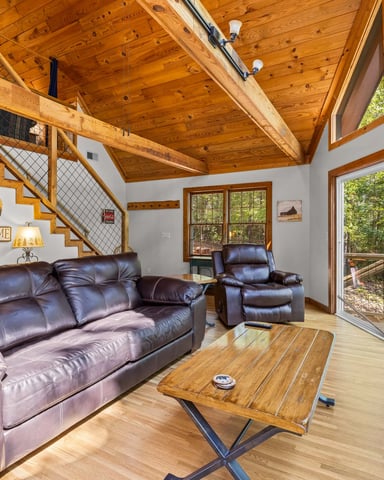 Cabin living room with exposed wood beams, leather seating, wooden coffee table, and glass doors opening to forest deck