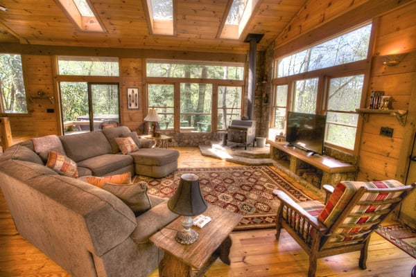 Spacious log cabin living room with wood beams, skylights, stone fireplace, gray sectional sofa, and expansive forest views through large windows.