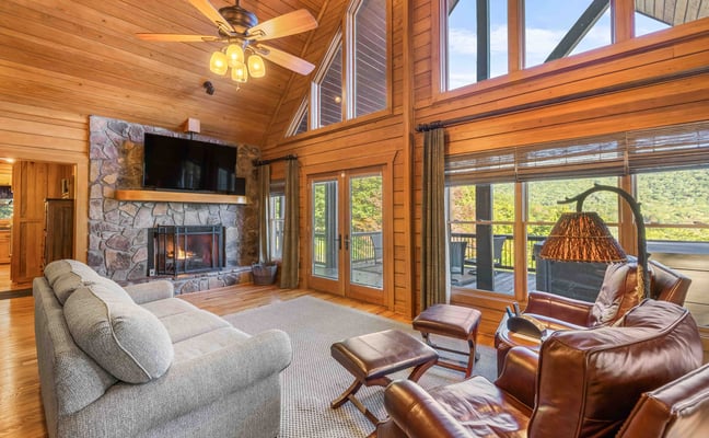 Spacious wood-paneled living room with stone fireplace, leather seating, ceiling fan, and expansive windows overlooking forested landscape.