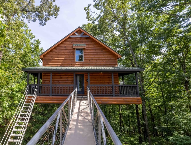Elevated log cabin with metal roof and wraparound porch, surrounded by dense green forest